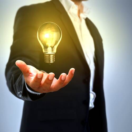 Mann im Anzug hält leuchtende Glühbirne schwebend über der offenen Hand vor grauem Hintergrund.