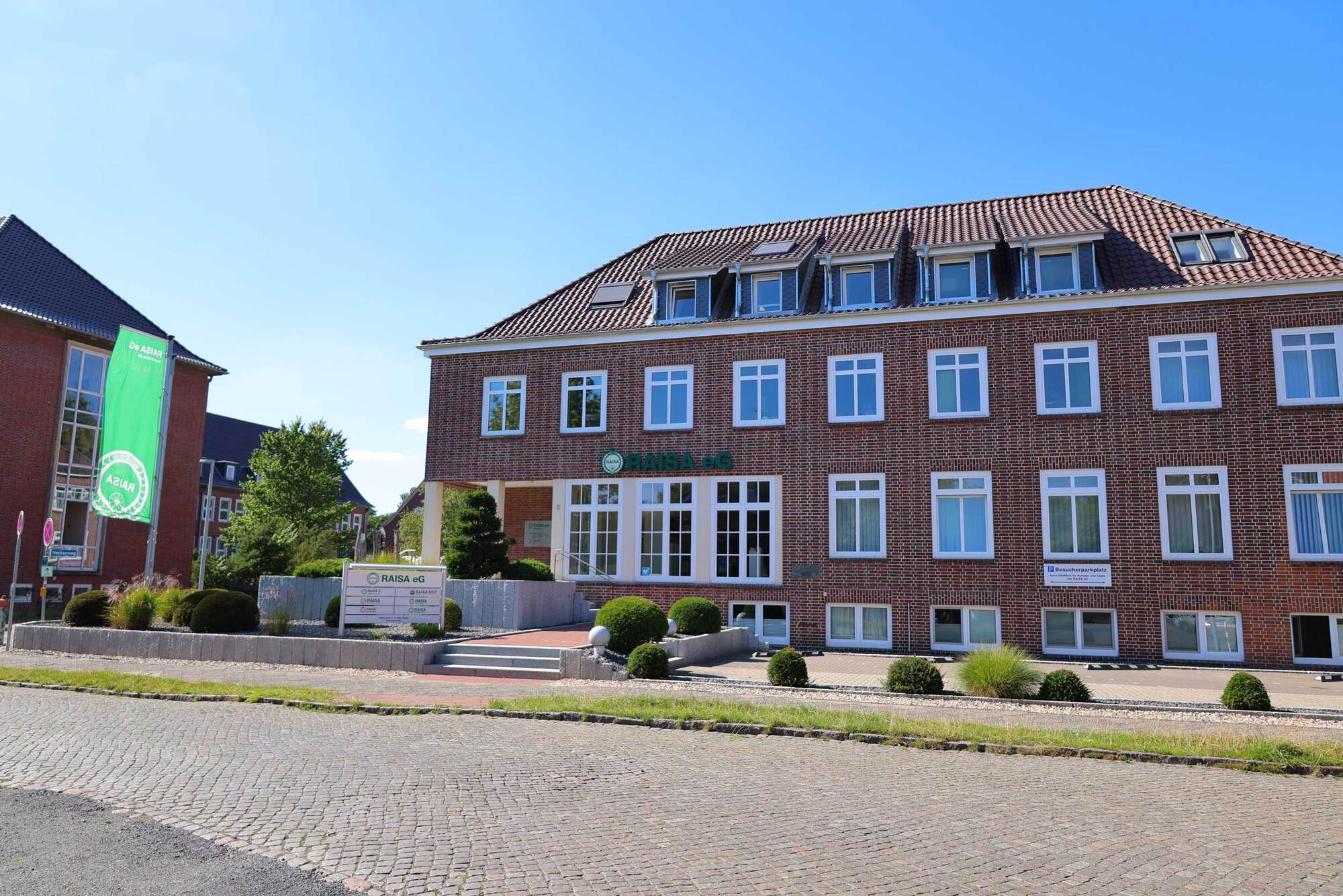 Backsteingebäude der RAISA eG mit gepflegtem Vorplatz und grünem Firmenschild bei Sonnenschein.