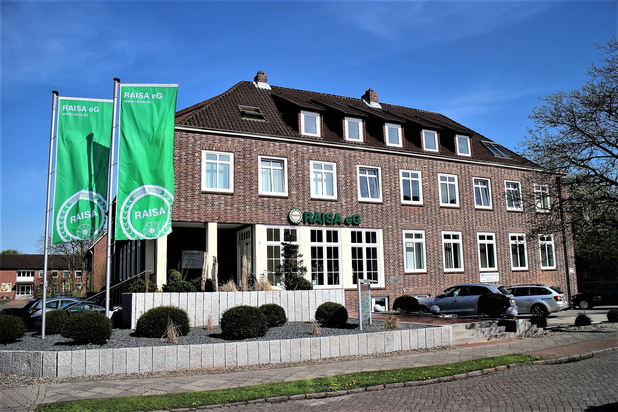 Backsteingebäude der RAISA eG mit gepflegtem Vorplatz und grünem Firmenschild bei Sonnenschein.
